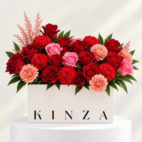 A floral arrangement in a white rectangular box with the brand name KINZA printed on the front. The bouquet features red and pink roses, red and pink carnations, and light pink ornamental grass, set against a clean white background on a white pedestal.