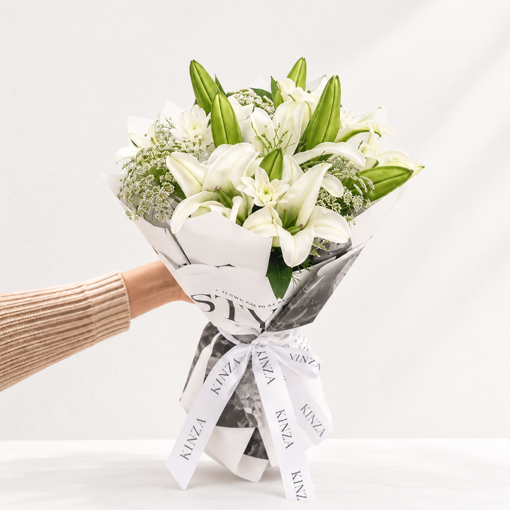 A bouquet of white lilies and green foliage wrapped in white and gray patterned paper with a white ribbon printed with the brand name 'KINZA', held by a person's hand wearing a beige ribbed sweater against a clean white background.