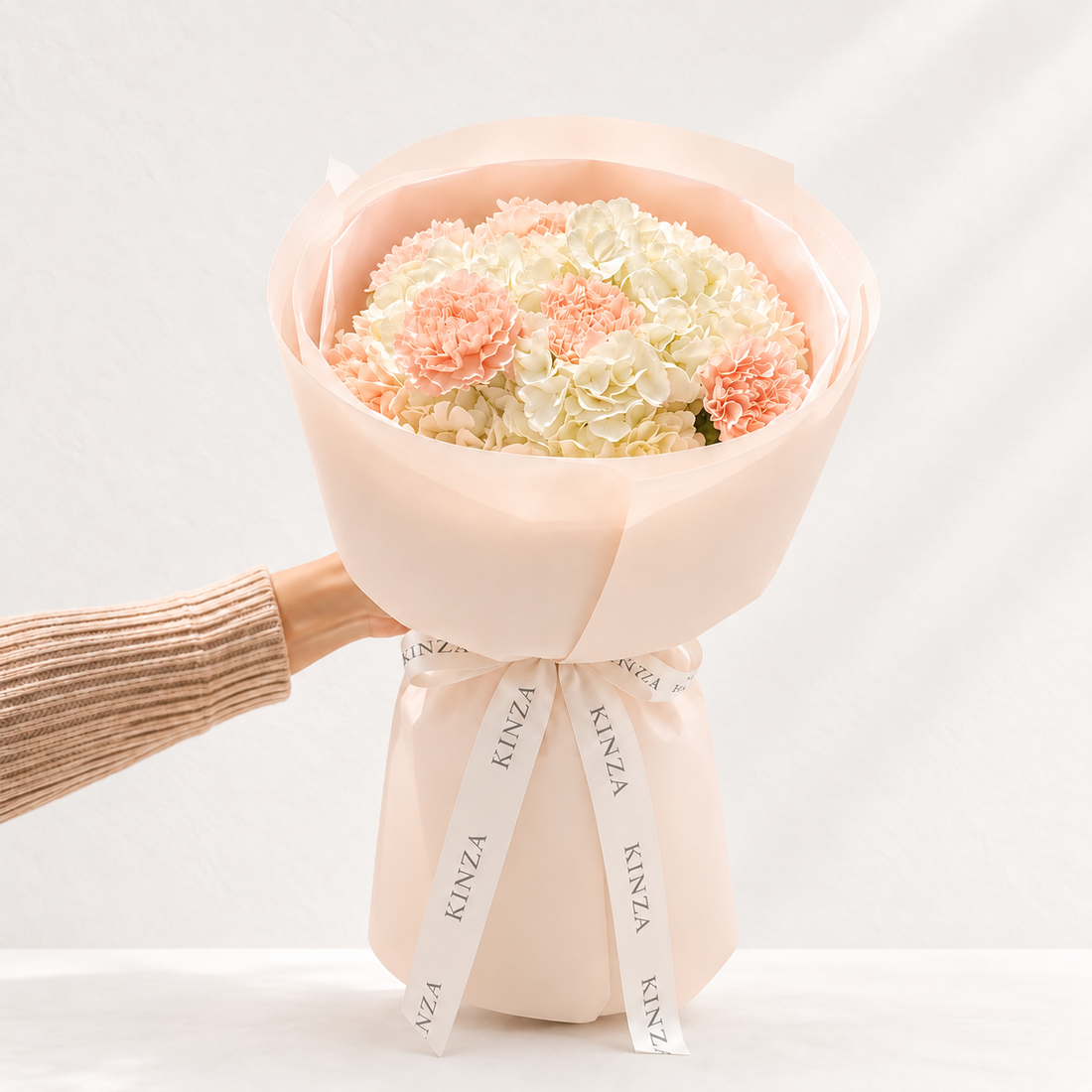 A bouquet of white and pink carnations wrapped in pale blush pink paper, tied with a white ribbon printed with the brand name KINZA, held by a person's hand wearing a beige ribbed sweater against a clean white background.