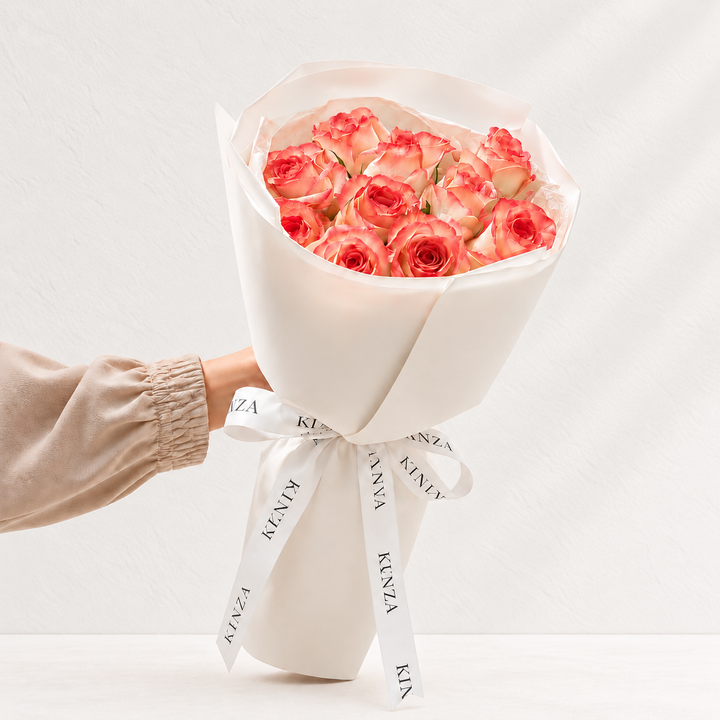 A bouquet of pink roses wrapped in white paper and tied with a white ribbon printed with the brand name KINZ, held by a person's hand against a clean white background.