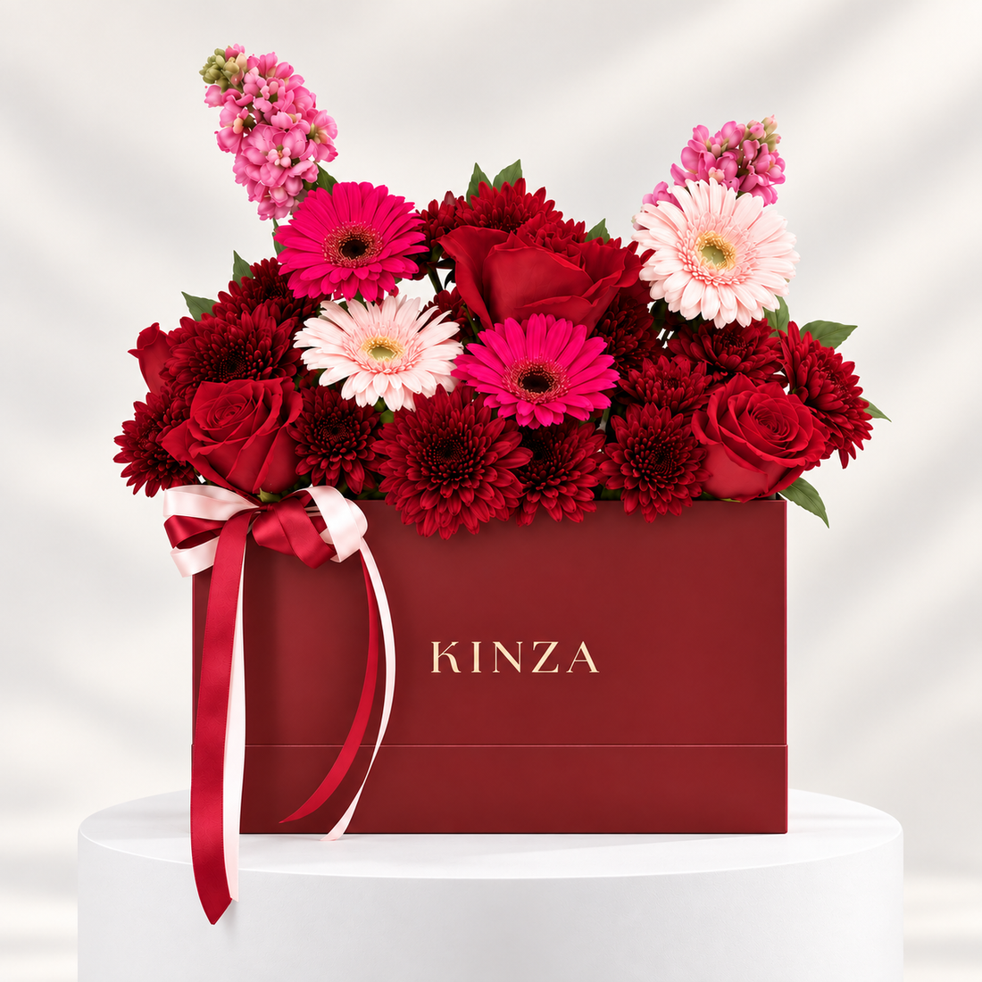 A floral gift box arrangement with red and pink flowers in a dark red KINZA box, tied with a red and white ribbon, displayed on a white pedestal against a light gray background.