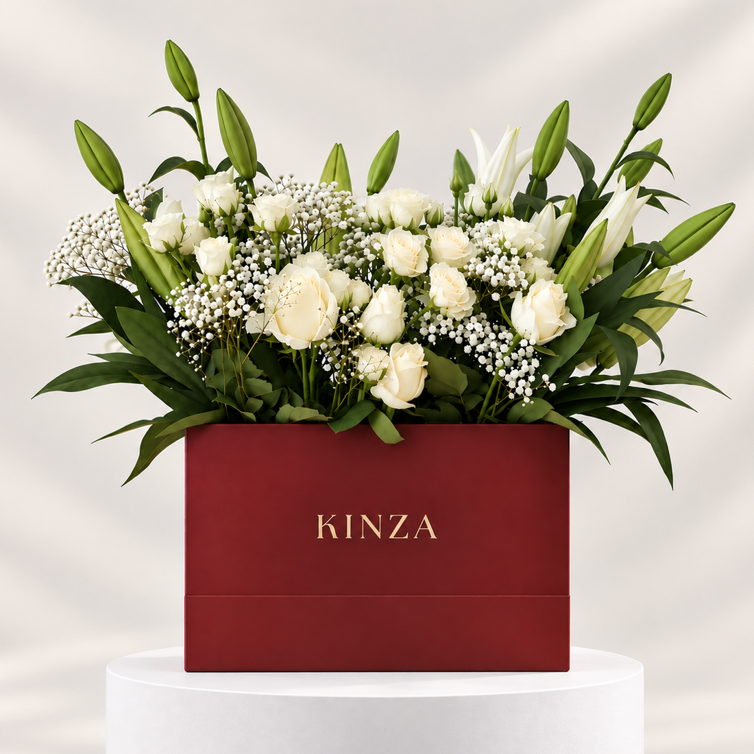 A floral arrangement in a red rectangular box with the brand name KINZA in gold lettering, displayed on a white pedestal against a light gray background. The bouquet features white roses, lilies, green leaves, and baby's breath.