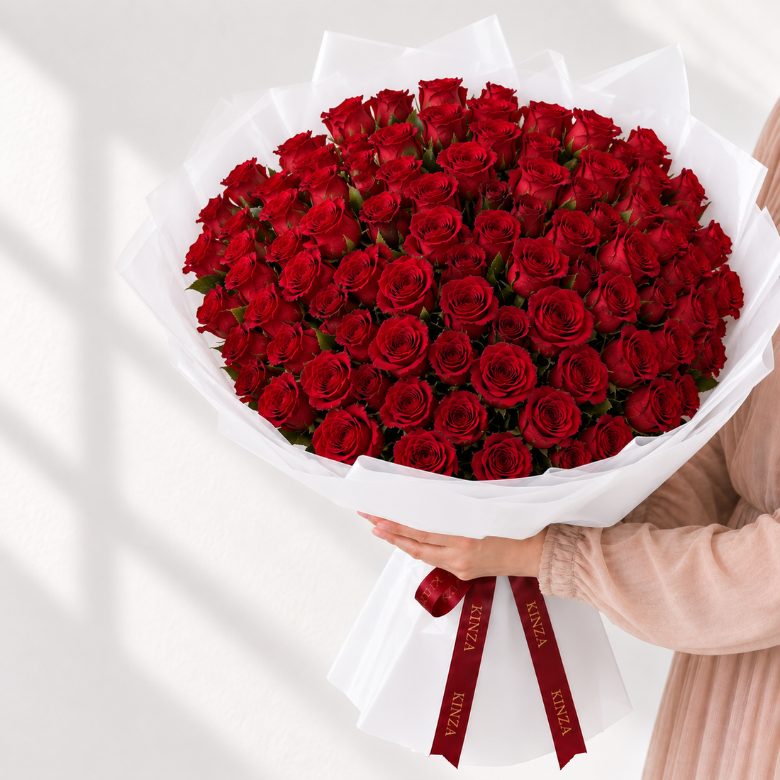 A large bouquet of red roses wrapped in white paper and tied with a red ribbon, held by a person wearing a light pink top against a bright white background.