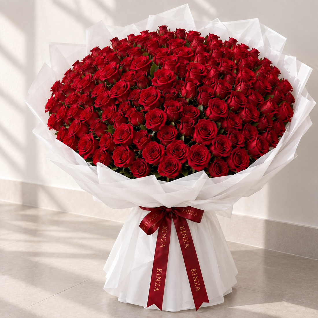 A large bouquet of red roses wrapped in white tissue paper and tied with a red ribbon printed with the brand name 'ANZA', set against a neutral indoor background.