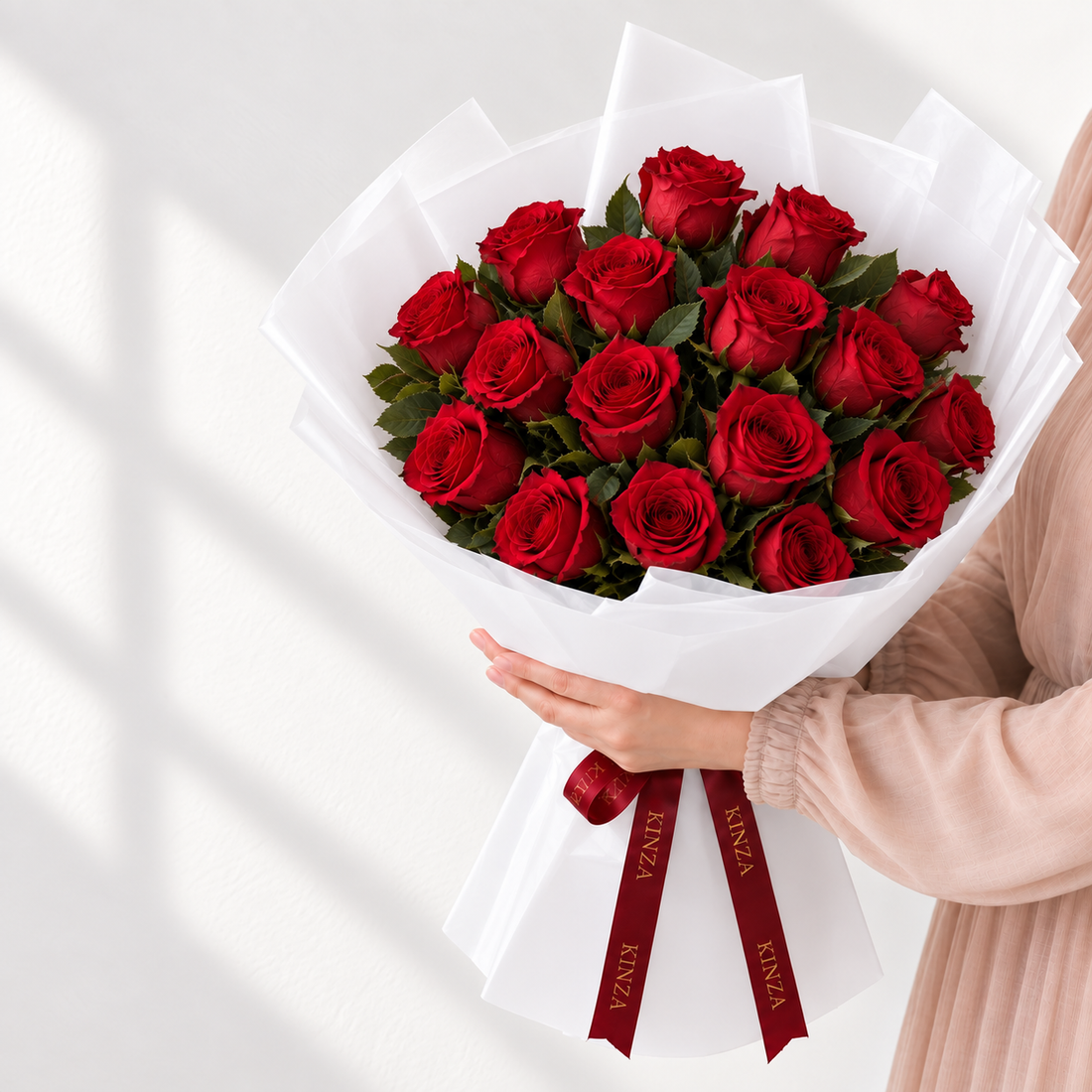 A bouquet of 20 red roses wrapped in white tissue paper with a dark red ribbon printed with the brand name 'KINZA', held by a person wearing a light pink top against a white background with soft window shadows.