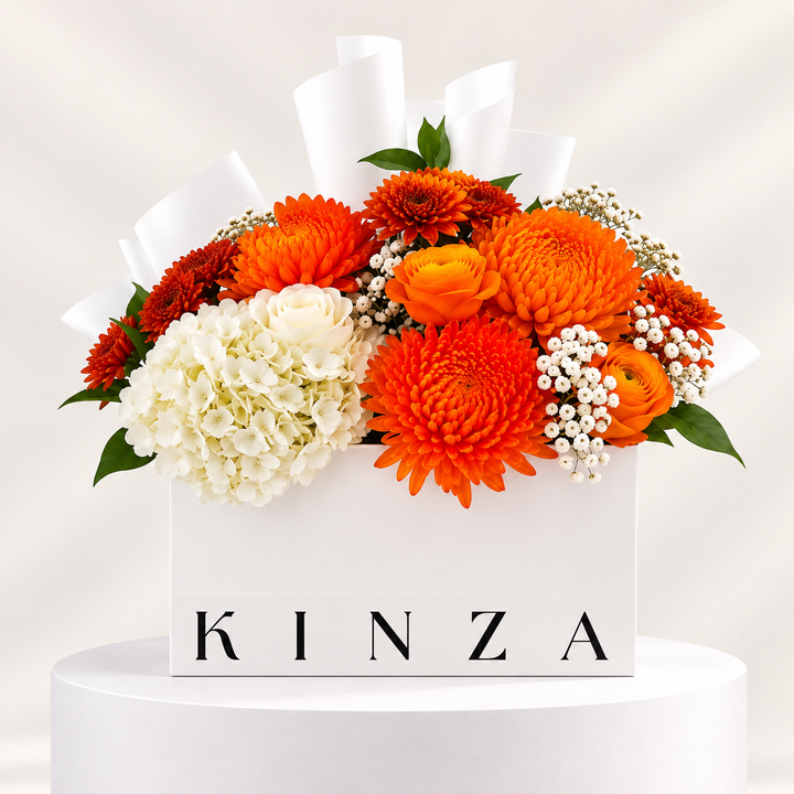 A white floral gift box with the brand name KINZA printed in black on the front, filled with a vibrant bouquet of orange and white flowers including chrysanthemums, roses, hydrangeas, and baby's breath, set on a white pedestal against a soft light background.
