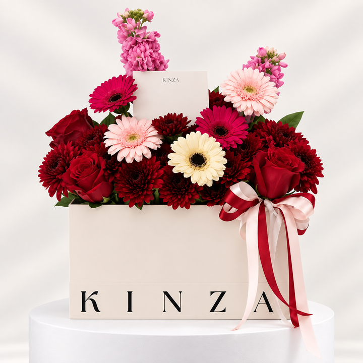 A floral gift box arrangement from KINZA, featuring deep red and pink roses, pink and cream gerbera daisies, and pink stock flowers, tied with a red and pale pink ribbon and presented on a white pedestal.