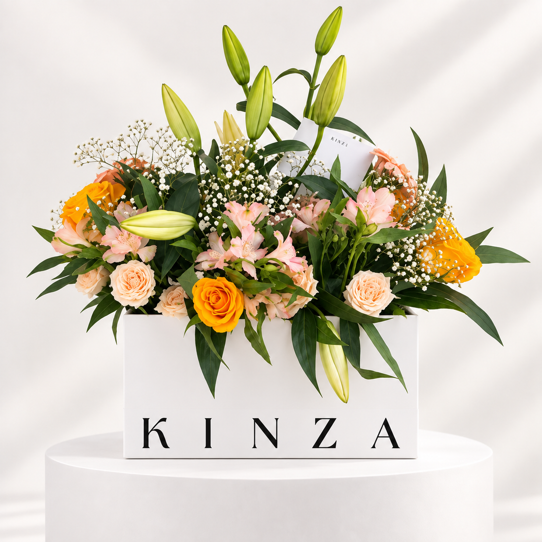 A floral arrangement in a white rectangular box with the brand name KINZA printed on the front. The bouquet includes orange roses, pale pink roses, lily buds, alstroemeria, baby's breath, and green foliage, displayed on a white pedestal against a white background with soft shadow overlays.