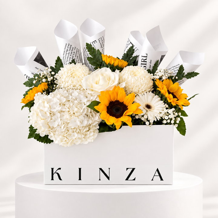 A floral arrangement in a white rectangular box with the name KINZA printed on the front, displayed on a white pedestal. The bouquet includes white hydrangeas, white roses, yellow sunflowers, white daisies, green foliage, and rolled newspaper-style accents rising behind the flowers.