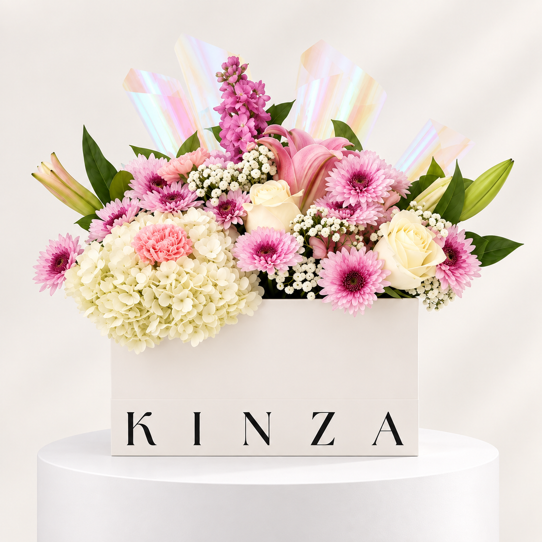 A floral arrangement in a white rectangular box with the brand name KINZA printed on the front, displayed on a white pedestal against a light neutral background. The bouquet features pink and white flowers, including chrysanthemums, roses, carnations, and hydrangeas, accented with iridescent paper strips.