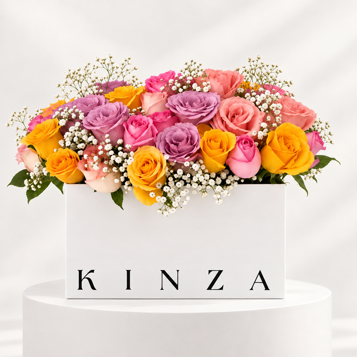 A vibrant floral arrangement of pink, purple, orange, and yellow roses with white baby's breath, displayed in a white rectangular box with the brand name 'KINZA' printed on the front, set on a white pedestal against a light gray background.