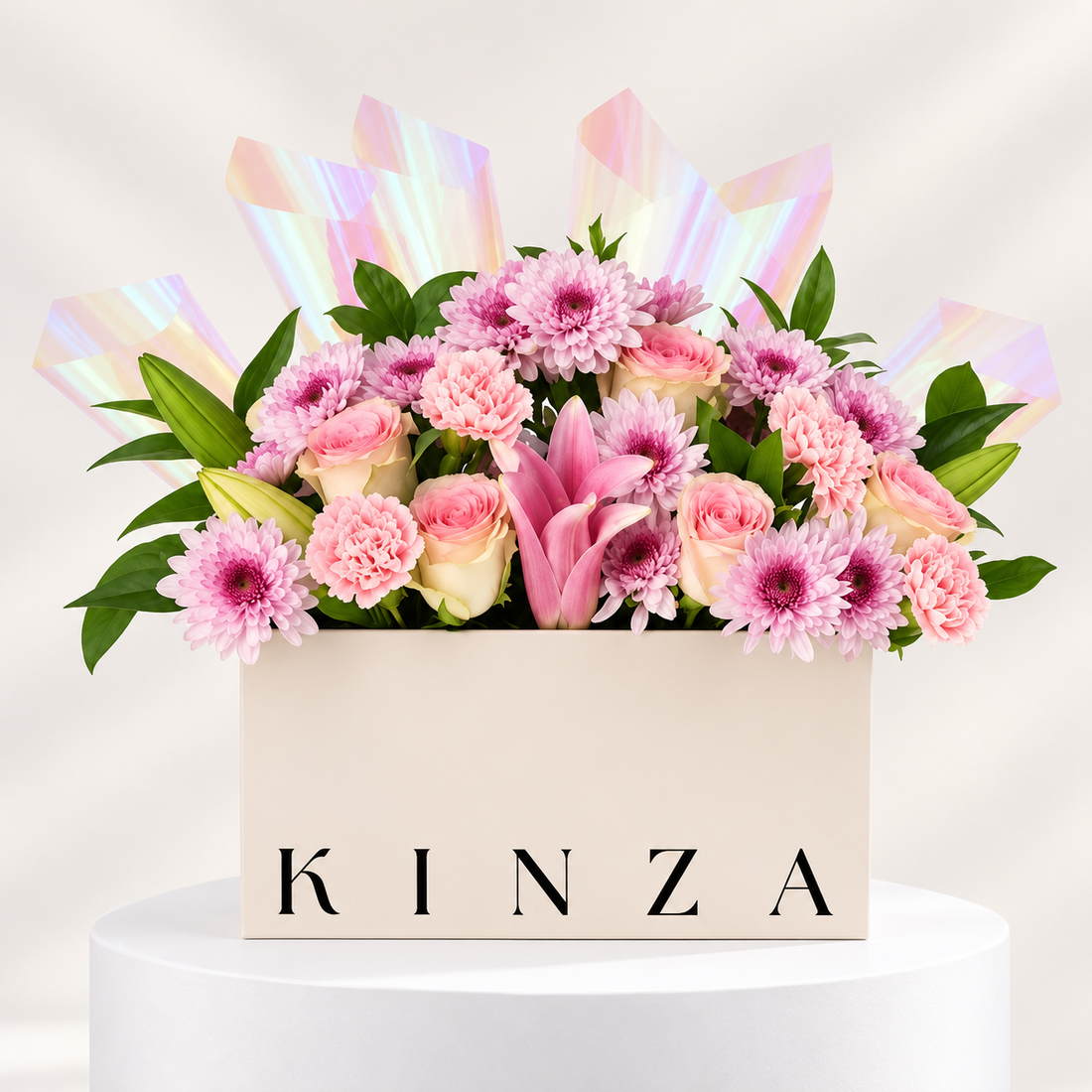 A floral arrangement in a cream-colored rectangular box with the brand name 'KINZA' printed on the front. The bouquet features pink and white flowers, including daisies, carnations, roses, and a pink lily, accented with iridescent pink wrapping paper, set on a white pedestal against a light beige background.