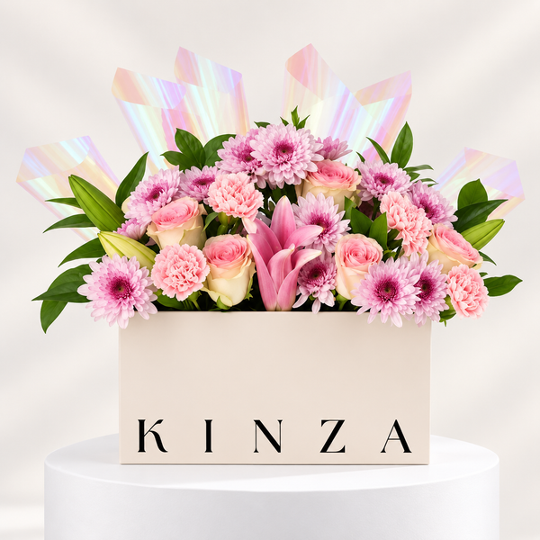 A floral arrangement in a cream-colored rectangular box with the brand name 'KINZA' printed on the front. The bouquet features pink and white flowers, including daisies, carnations, roses, and a pink lily, accented with iridescent pink wrapping paper, set on a white pedestal against a light beige background.