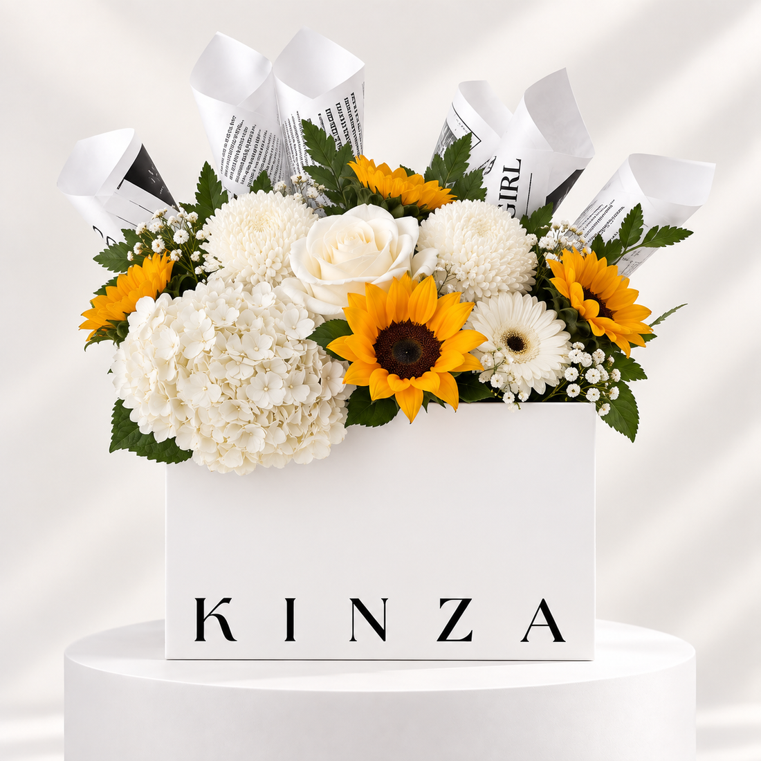 A white rectangular flower box with the brand name KINZA printed in black on the front, filled with a bouquet of white hydrangeas, white roses, yellow sunflowers, white daisies, green leaves, and rolled newspaper stems, displayed on a white pedestal against a light draped background.