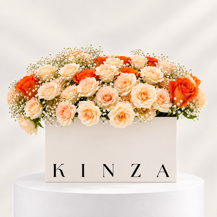 A floral arrangement of peach, cream, and orange roses with baby's breath in a white rectangular box printed with the brand name 'KINZA', displayed on a white pedestal against a soft light gray background.