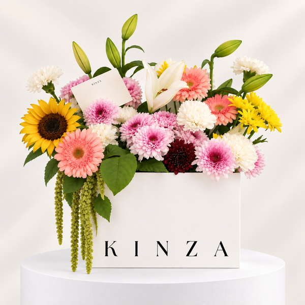 A floral arrangement in a white rectangular box printed with the name KINZA, featuring pink, yellow, white, and burgundy flowers with green leaves and buds, displayed on a white pedestal against a soft light background.