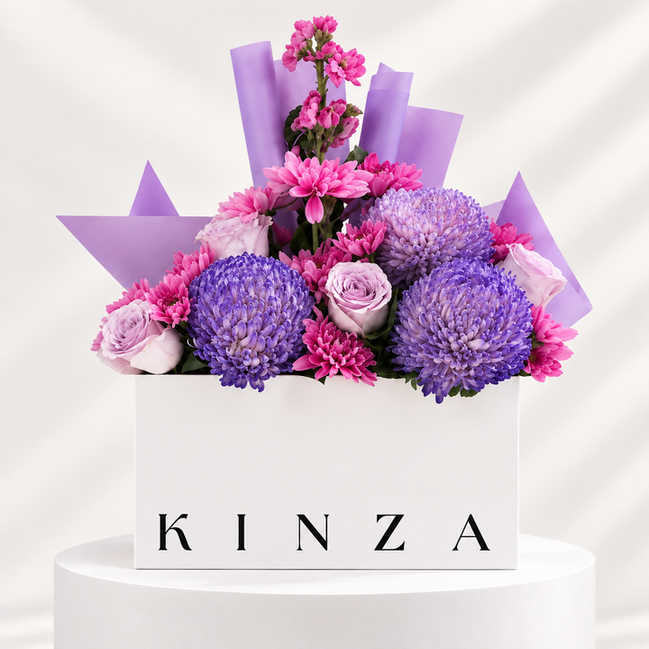 A floral arrangement in a white rectangular box with the brand name KINZA printed in black on the front. The bouquet features purple and pink flowers, including large purple chrysanthemums, pink carnations, and light purple roses, wrapped in lavender paper and set on a white pedestal against a light background.