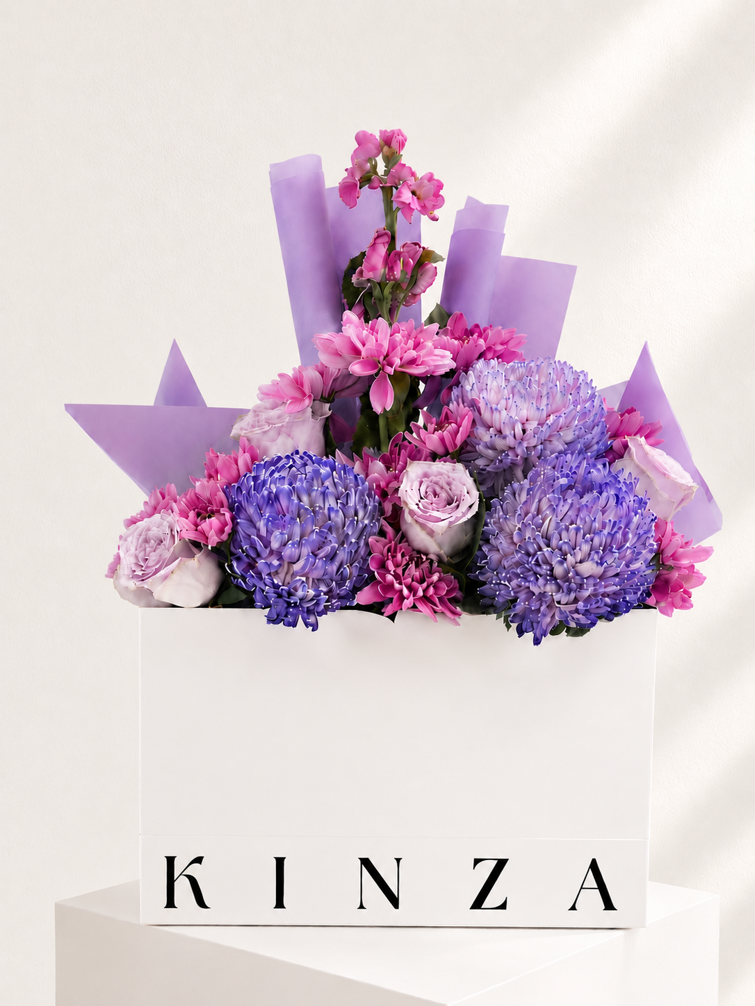 A floral arrangement in a white rectangular box with the brand name KINZA printed on the front. The bouquet features purple and pink flowers, including two large purple globes, pink chrysanthemums, and lavender roses, wrapped in light purple paper against a clean white background.