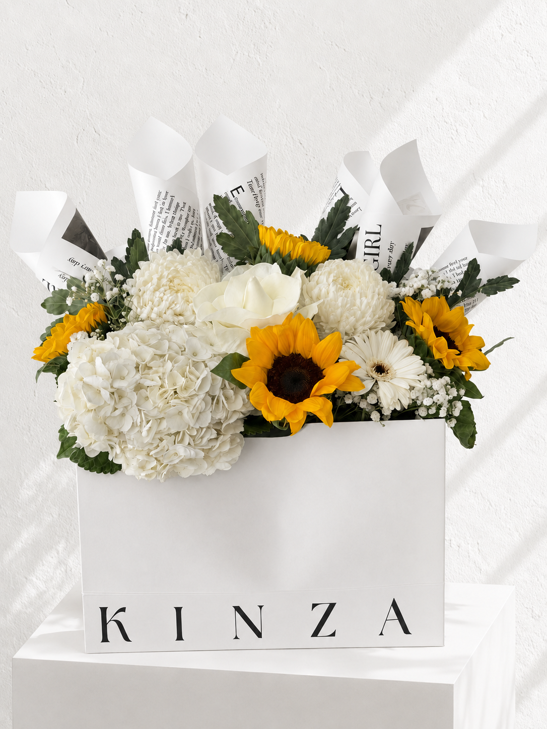 A floral arrangement in a white rectangular box with the brand name KINZA printed on the front. The bouquet features white hydrangeas, white roses, yellow sunflowers, white daisies, and green foliage, wrapped with white paper printed with black text, set against a white textured wall.