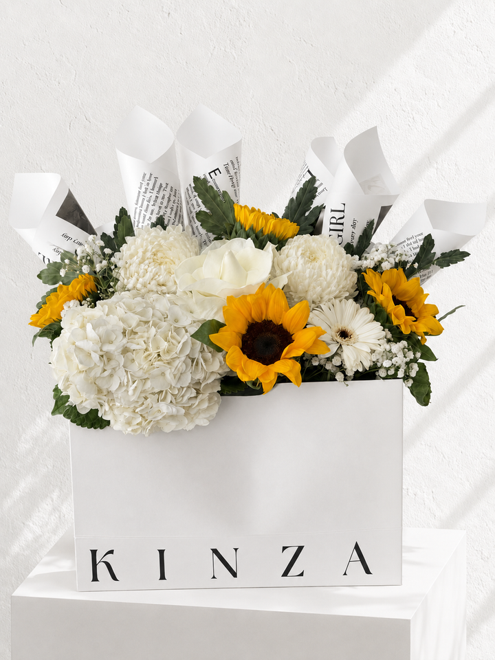 A floral arrangement in a white rectangular box with the brand name KINZA printed on the front. The bouquet features white hydrangeas, white roses, yellow sunflowers, white daisies, and green foliage, wrapped with white paper printed with black text, set against a white textured wall.