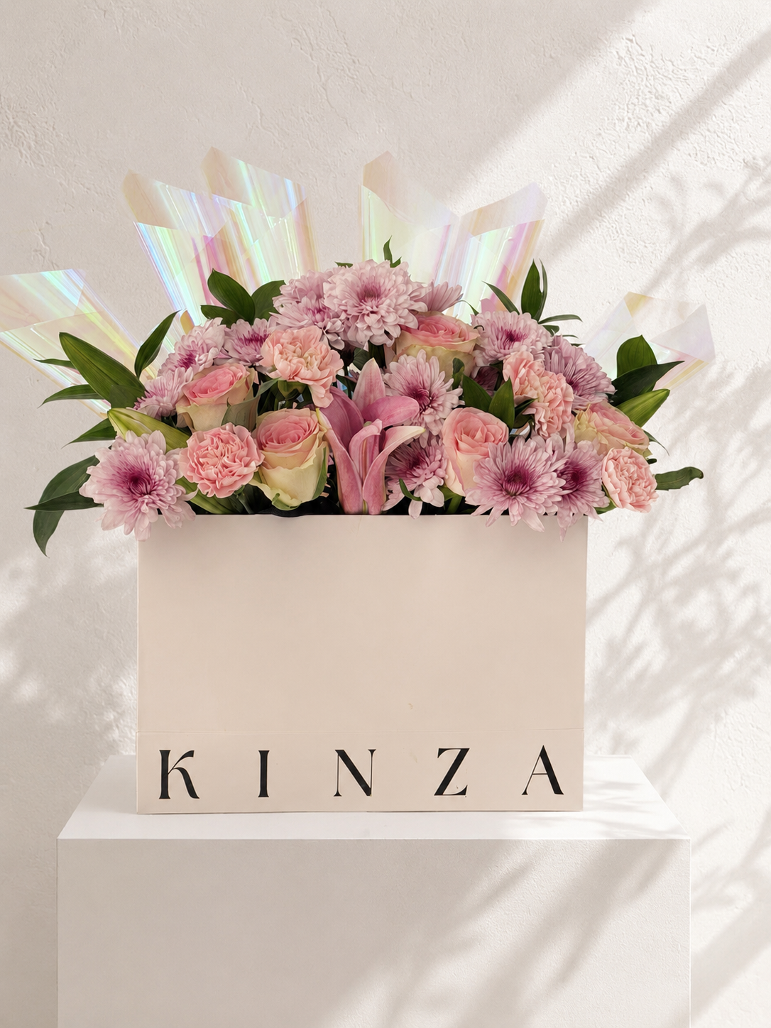 A floral arrangement in a white square box printed with the name KINZA, filled with pink and purple flowers, green leaves, and iridescent wrapping, displayed on a white pedestal against a textured wall with soft shadows.